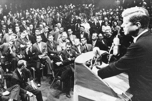 John F Kennedy in a black and white photo standing at a podium addressing a large crowd in a ballroom
