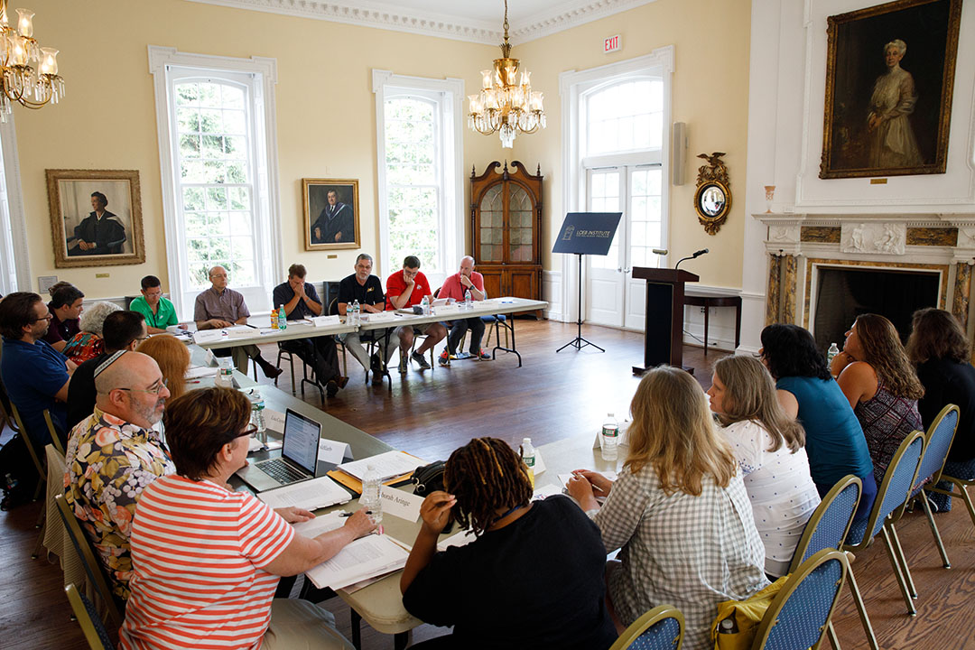 A large group of scholars seated at a horseshoe of tables having a discussion in a room with oil paintings and chandeliers